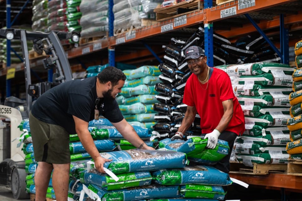 Two men stack bags of soil or fertilizer in a warehouse, smiling and working together. Shelves filled with more bags are visible in the background, and a forklift is seen to the left.