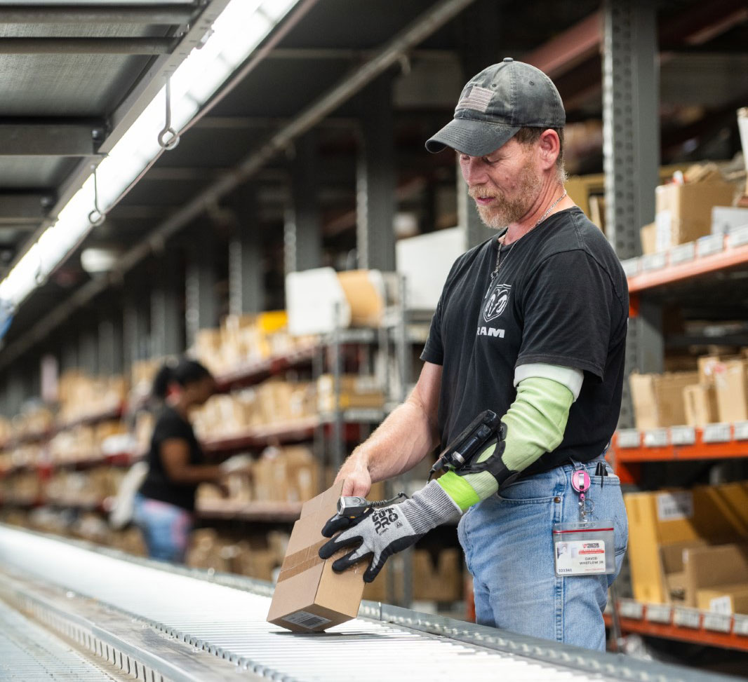 A man wearing a cap and gloves places a box on a conveyor belt in a warehouse, surrounded by shelves filled with packages. Another worker is visible in the background.