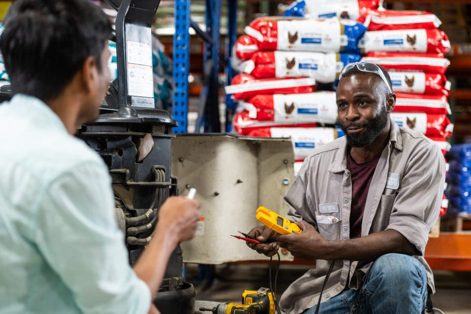 Two men work together in a warehouse. One, holding a multimeter and wire, kneels beside machinery and smiles. The other faces him, partially visible. Bags of pet food are stacked on shelves in the background.